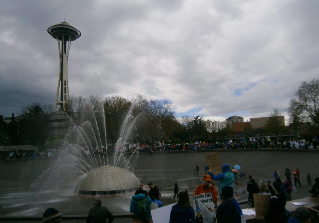 A crowd around the International Fountain at Seattle Center, with the Space Needle in the background, and the arches of the Pacific Science Center at right.